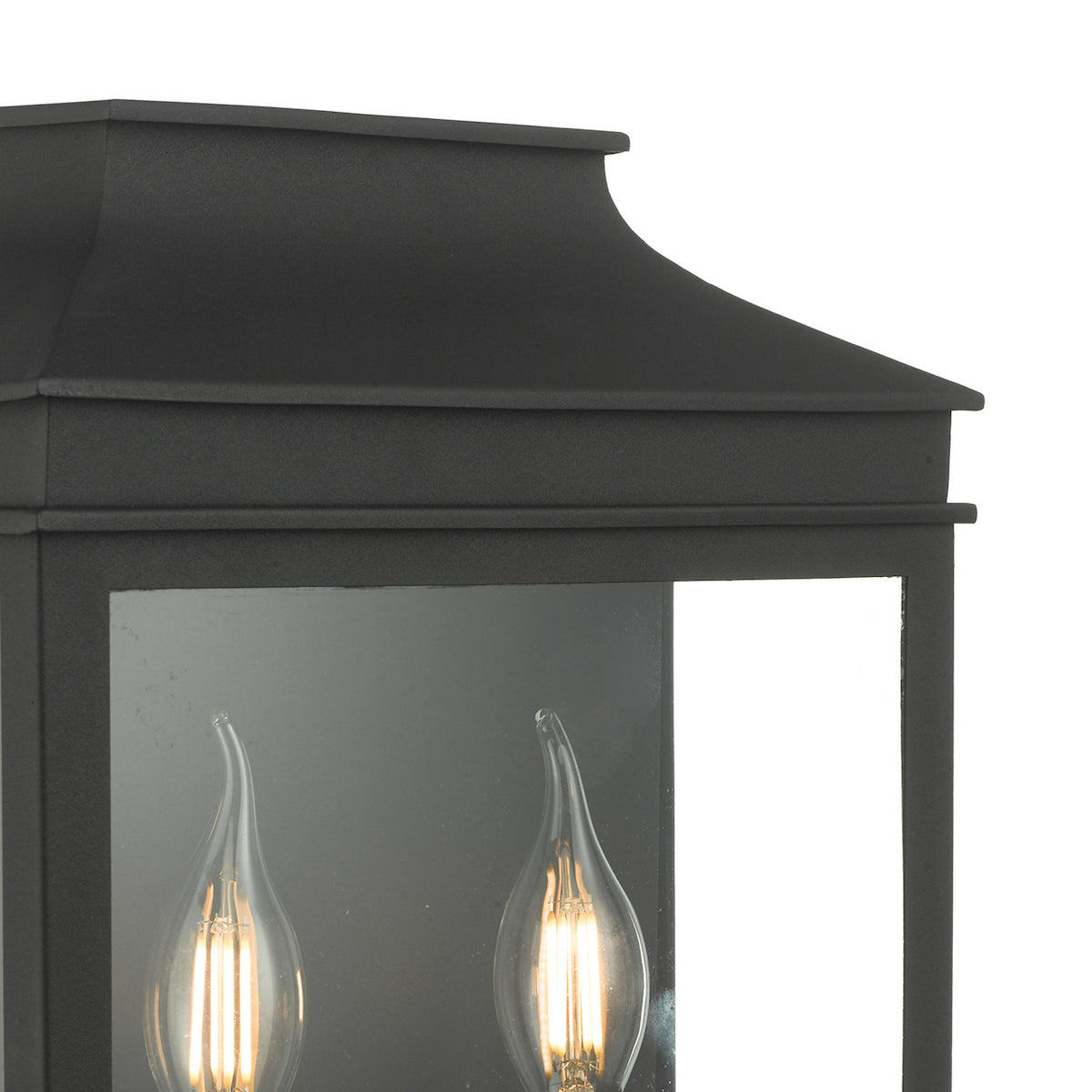 Close-up of a black outdoor wall lantern with two glowing, clear, flame-tip light bulbs inside.