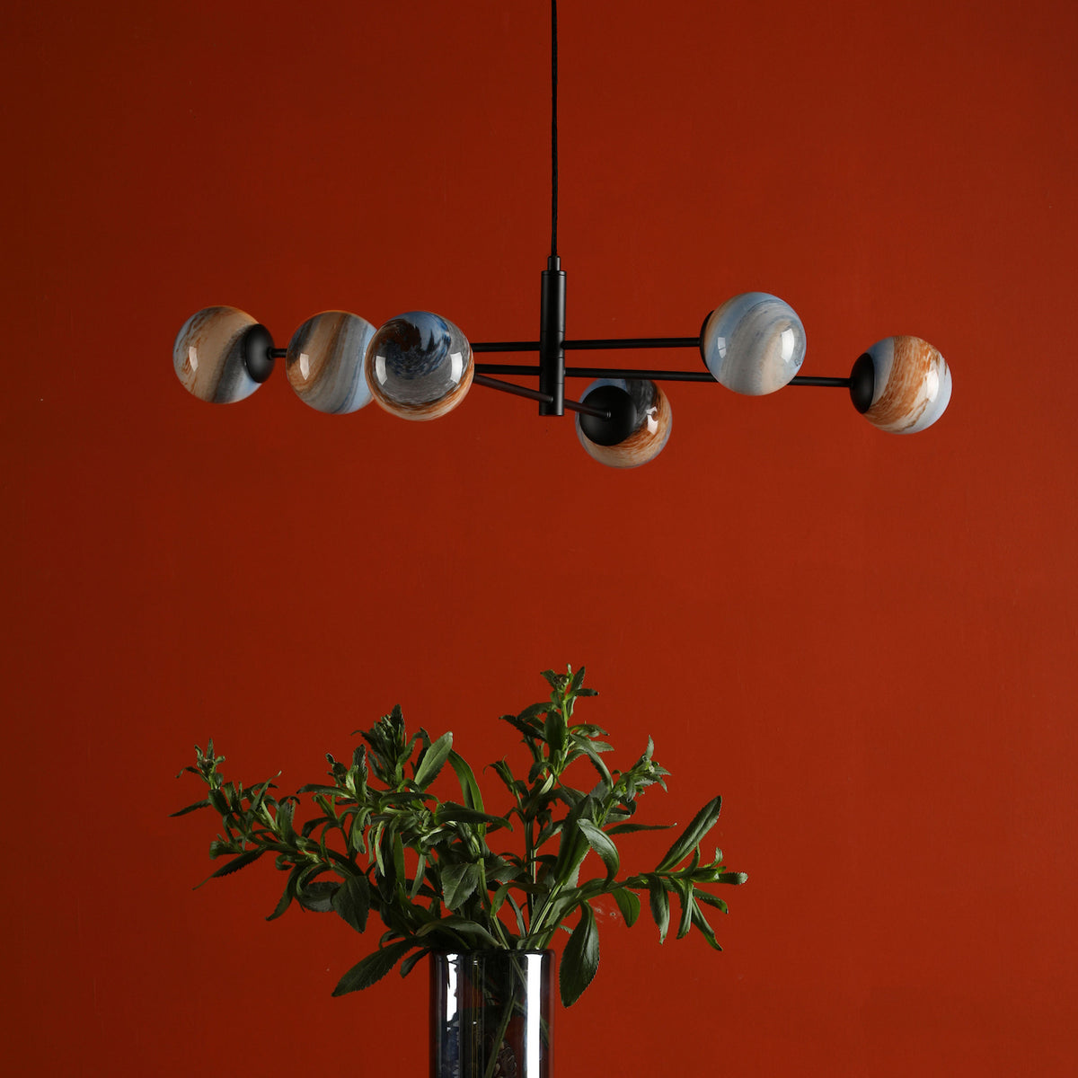 A modern chandelier with six spherical, marbled glass shades hangs above a vase of green foliage against a red wall.