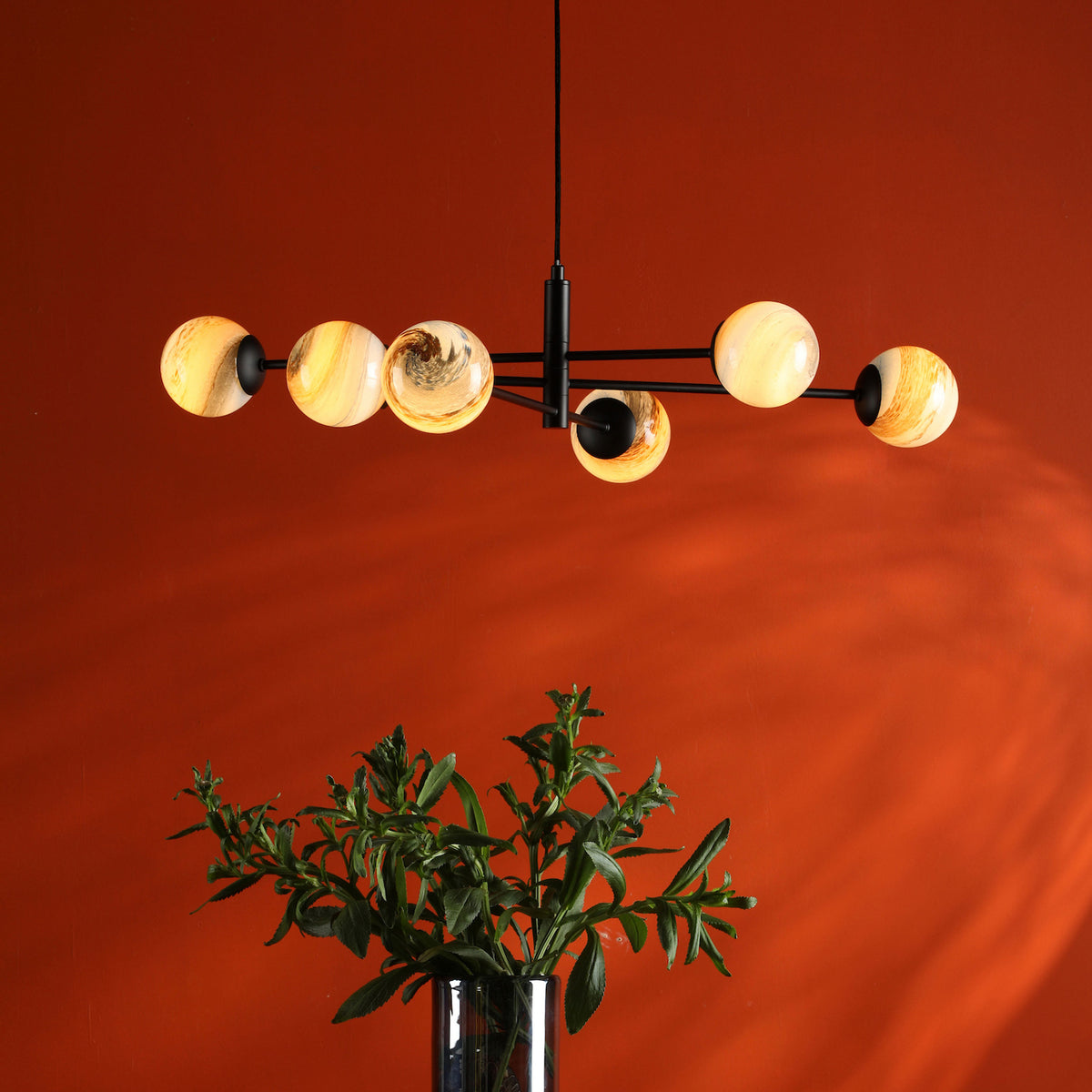 Modern chandelier with six amber globe lights, black frame, above a vase of green foliage against an orange wall.