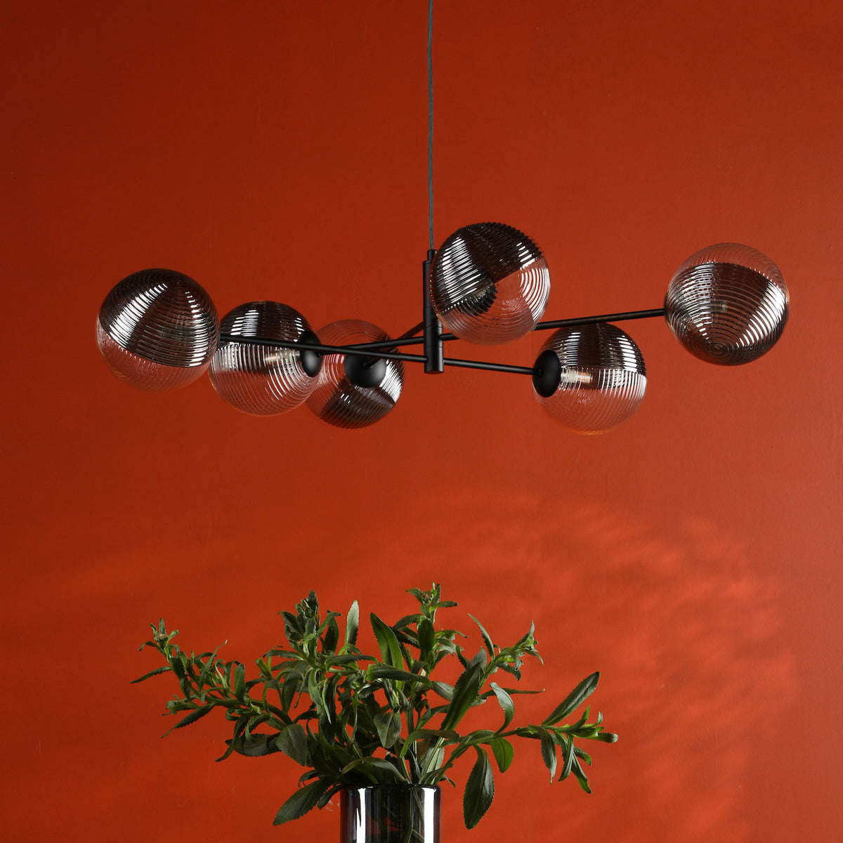 Modern black chandelier with six ribbed glass globes, above a vase of green foliage against an orange wall.