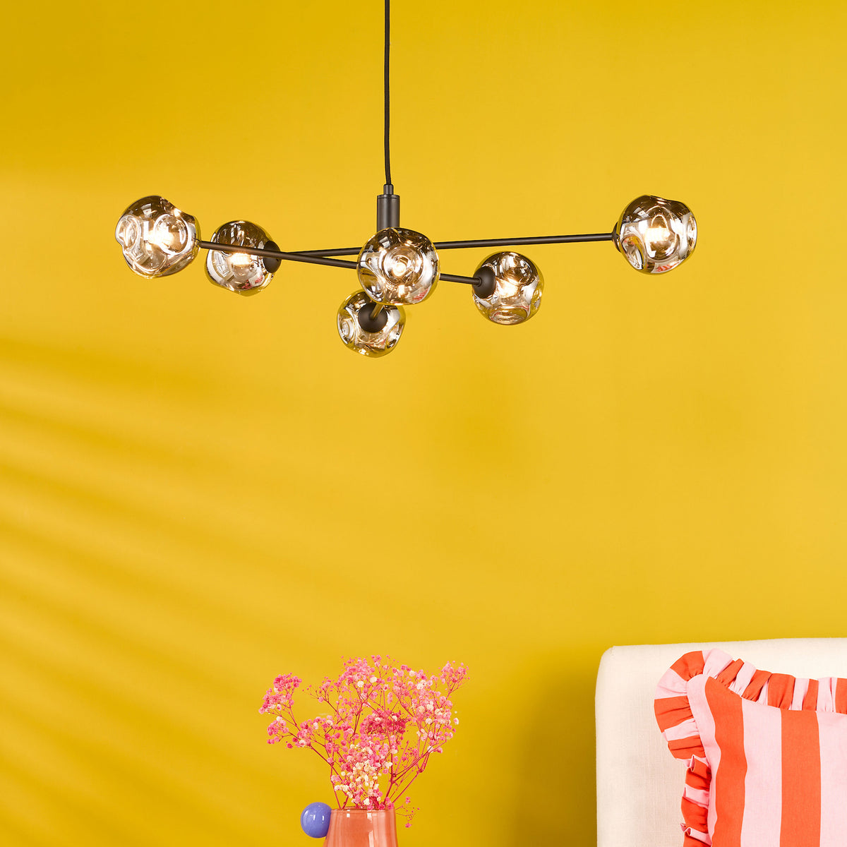 Modern chandelier with six glass globes on a black frame, above a vase of pink flowers and a striped pillow.