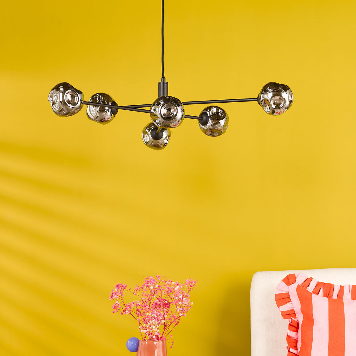Modern chandelier with six chrome-like globes on black rods, against a bright yellow wall. Pink flowers in a vase.