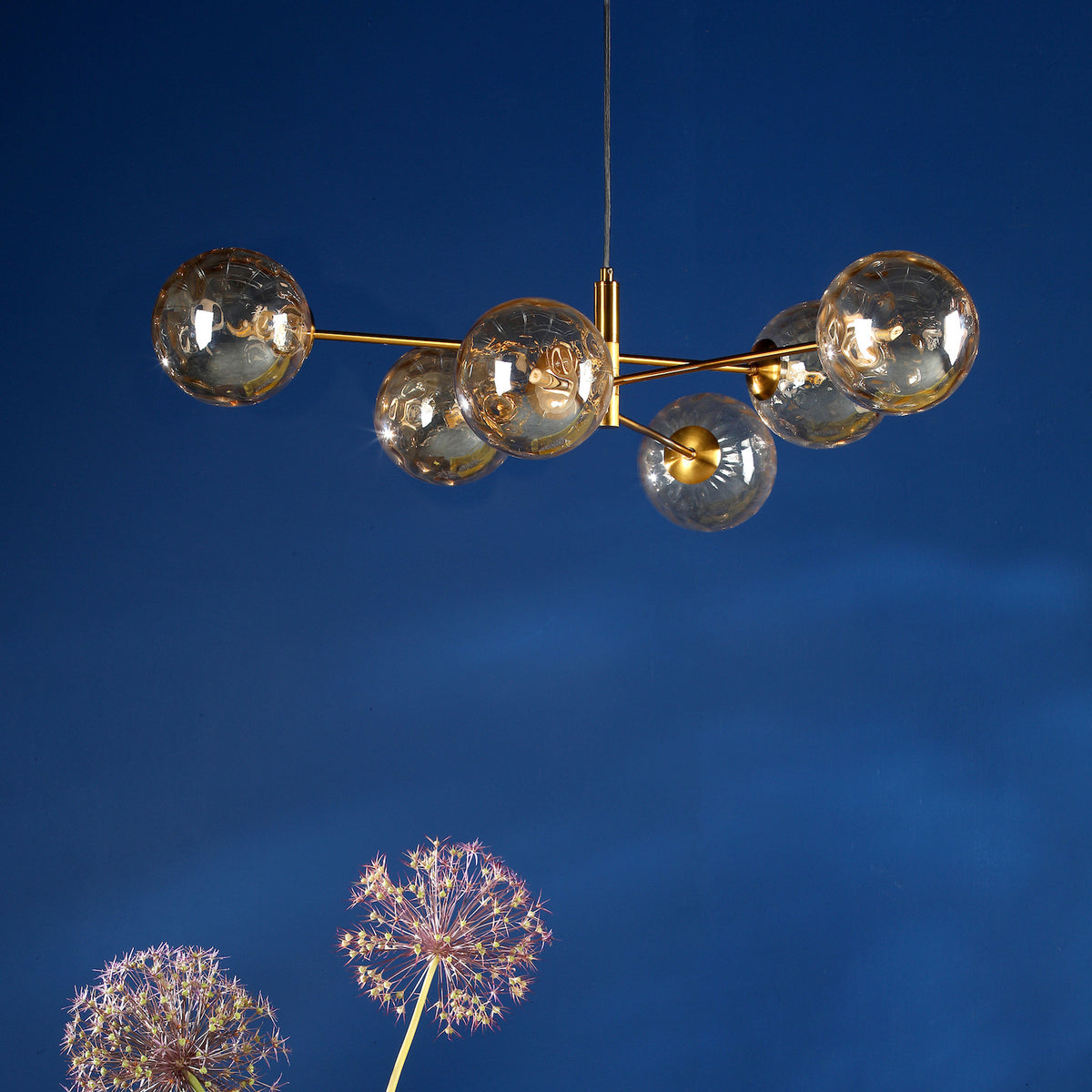 Modern gold chandelier with six clear glass globes, against a deep blue wall with two dandelion-like flowers.