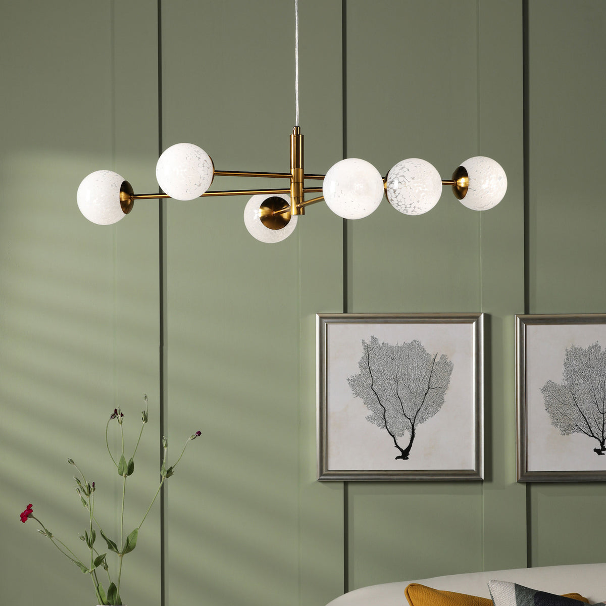 A modern gold chandelier with six white globe lights hangs from a ceiling, illuminating a green wall.