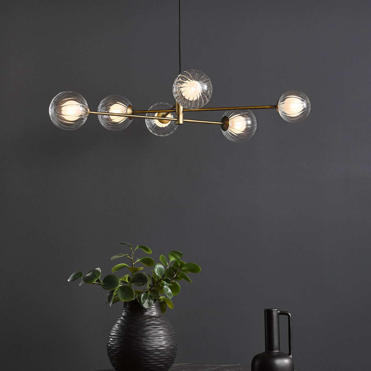 Modern chandelier with six clear, ribbed glass globes on brass arms. A black textured vase with greenery sits below.