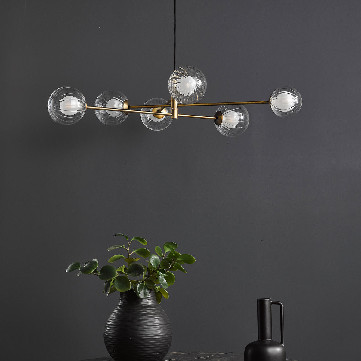 A modern brass chandelier with six clear, ribbed glass globes, illuminating a dark gray wall above a black vase with green foliage.