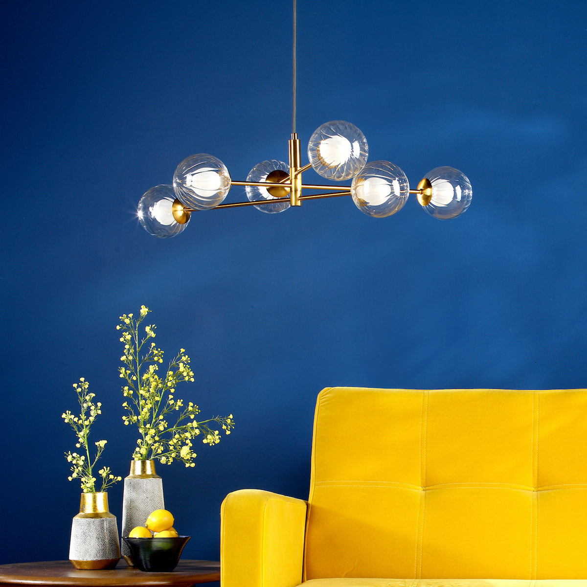 Modern gold chandelier with six clear globe lights hanging above a yellow sofa and vases with flowers.