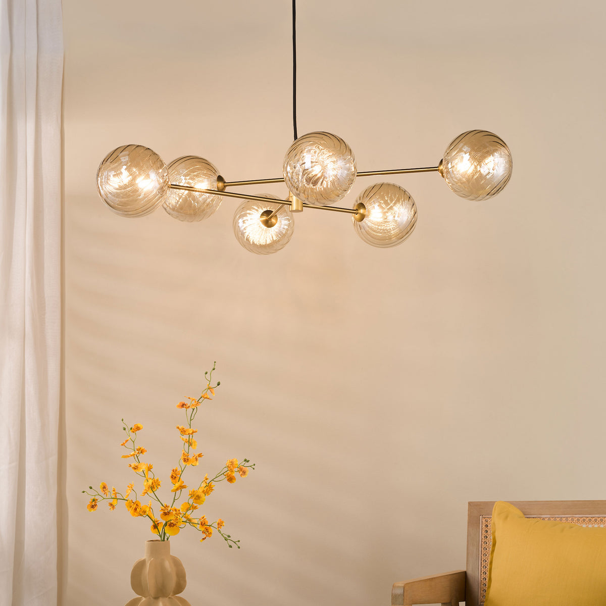 A modern chandelier with six amber glass globes, a vase of yellow flowers, and a wooden chair with a yellow cushion.