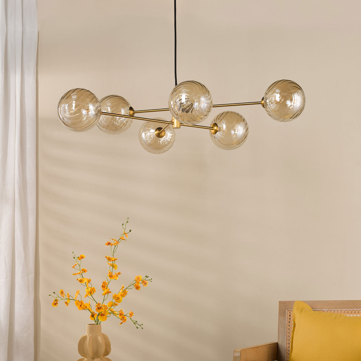 Modern chandelier with six amber glass globes, a vase of yellow flowers, and a wooden chair with a yellow pillow.