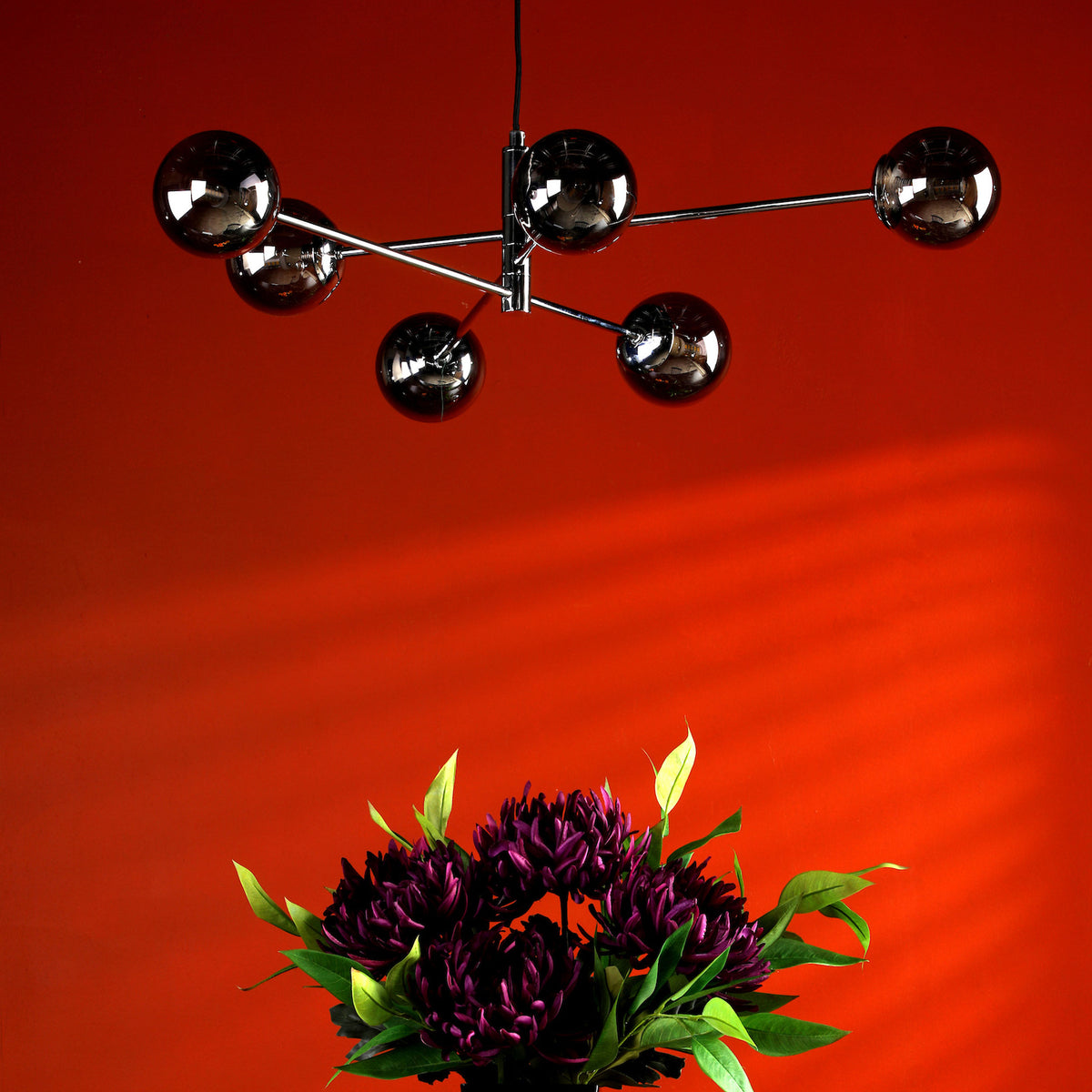 A modern chandelier with five spherical lights hangs above a bouquet of dark purple flowers against a red wall.