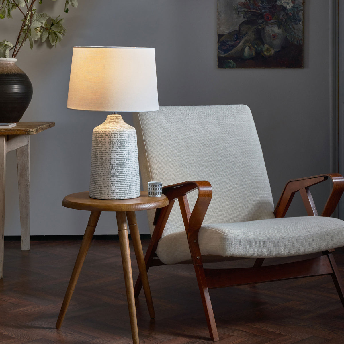 A mid-century modern armchair with a cream cushion and wooden frame next to a small wooden table with a lamp.