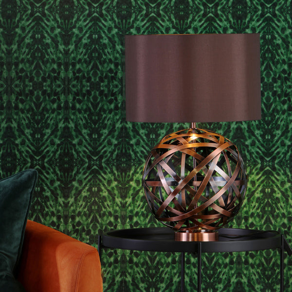 A bronze orb lamp with a dark shade sits on a black table next to an orange sofa, against green patterned wallpaper.