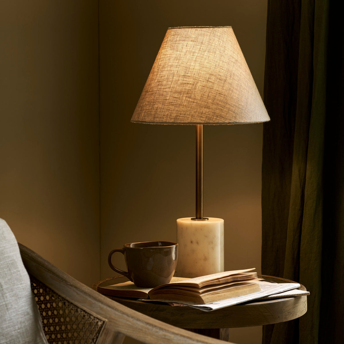 Winsley brass and marble table lamp with natural linen shade on a wooden table with a cup and books