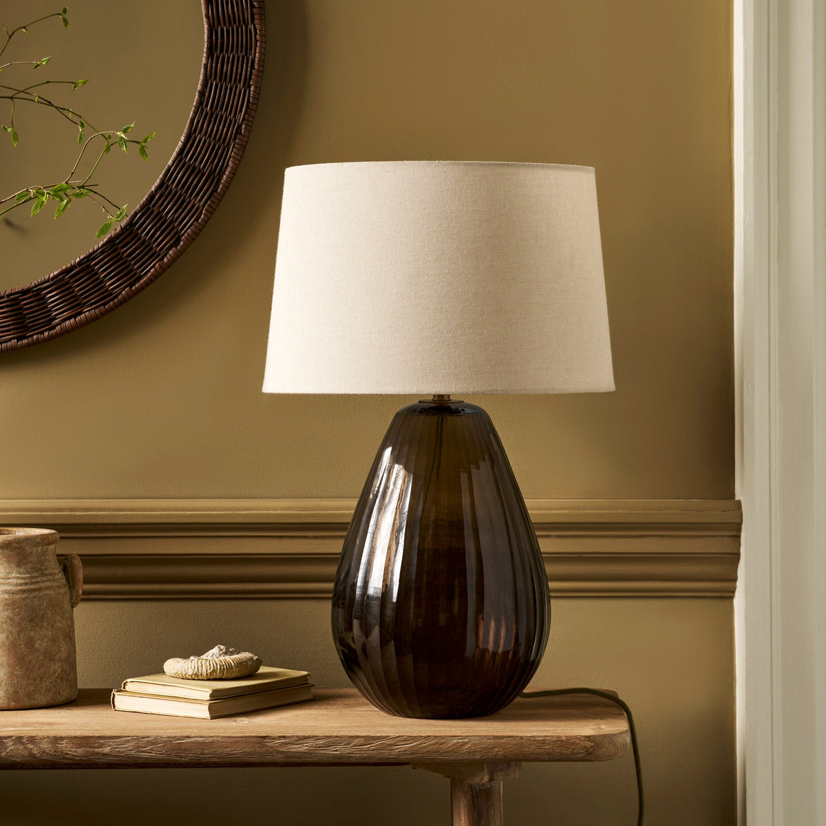 A table lamp with a ribbed dark amber glass base and a cream fabric drum shade, styled on a wooden console table against a warm brown wall.