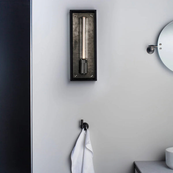 Black framed wall sconce with a cylindrical bulb, a black towel hook with a white towel, and a round mirror