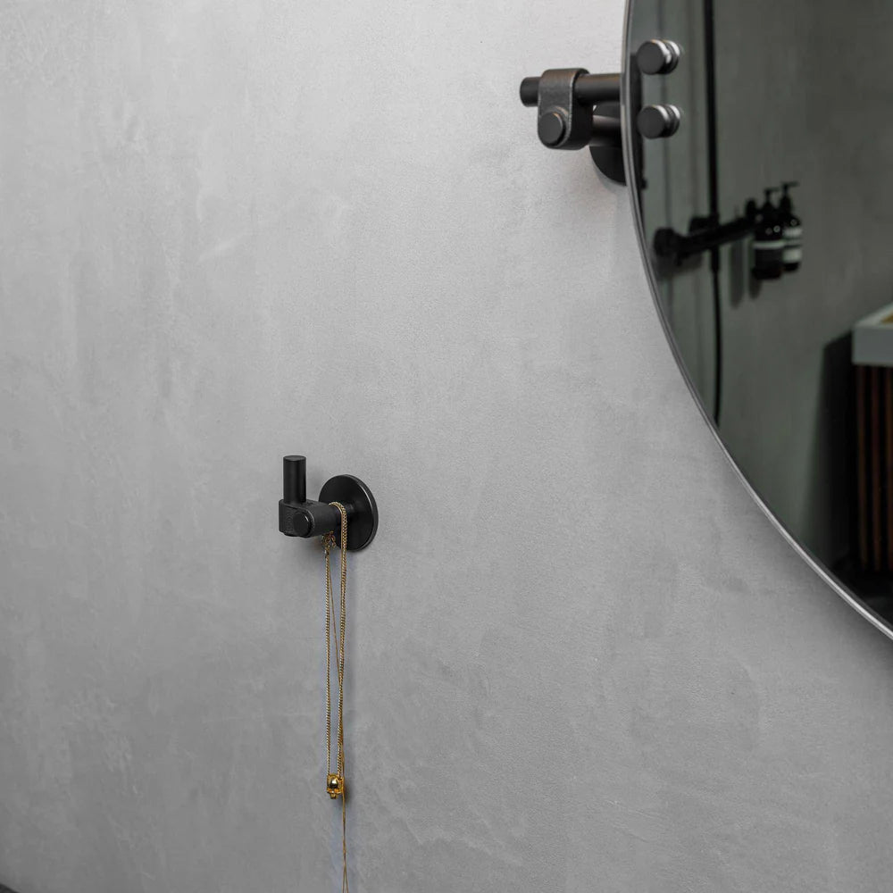Black wall hook with a gold necklace hanging on it, next to a round mirror