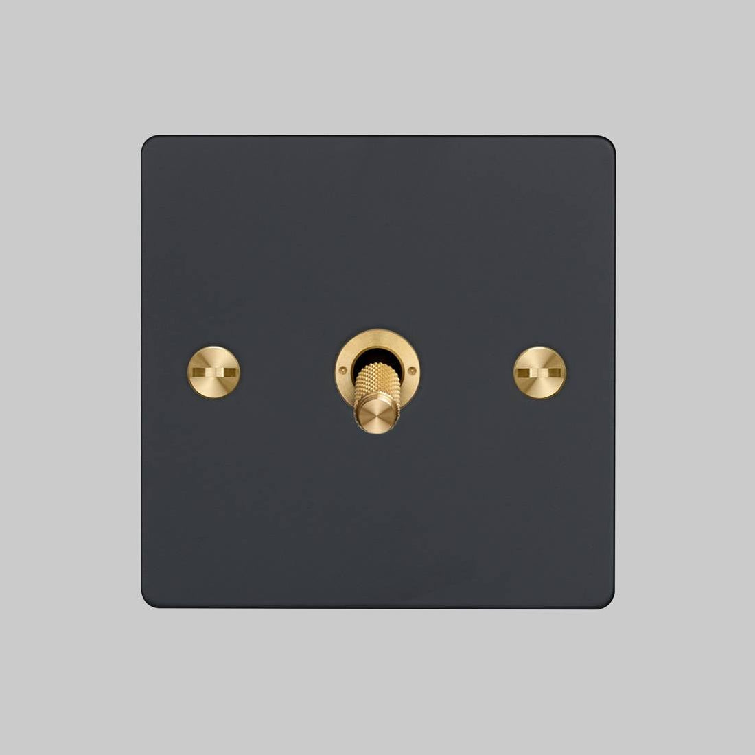 Black square light switch with a central brass toggle and two brass mounting screws