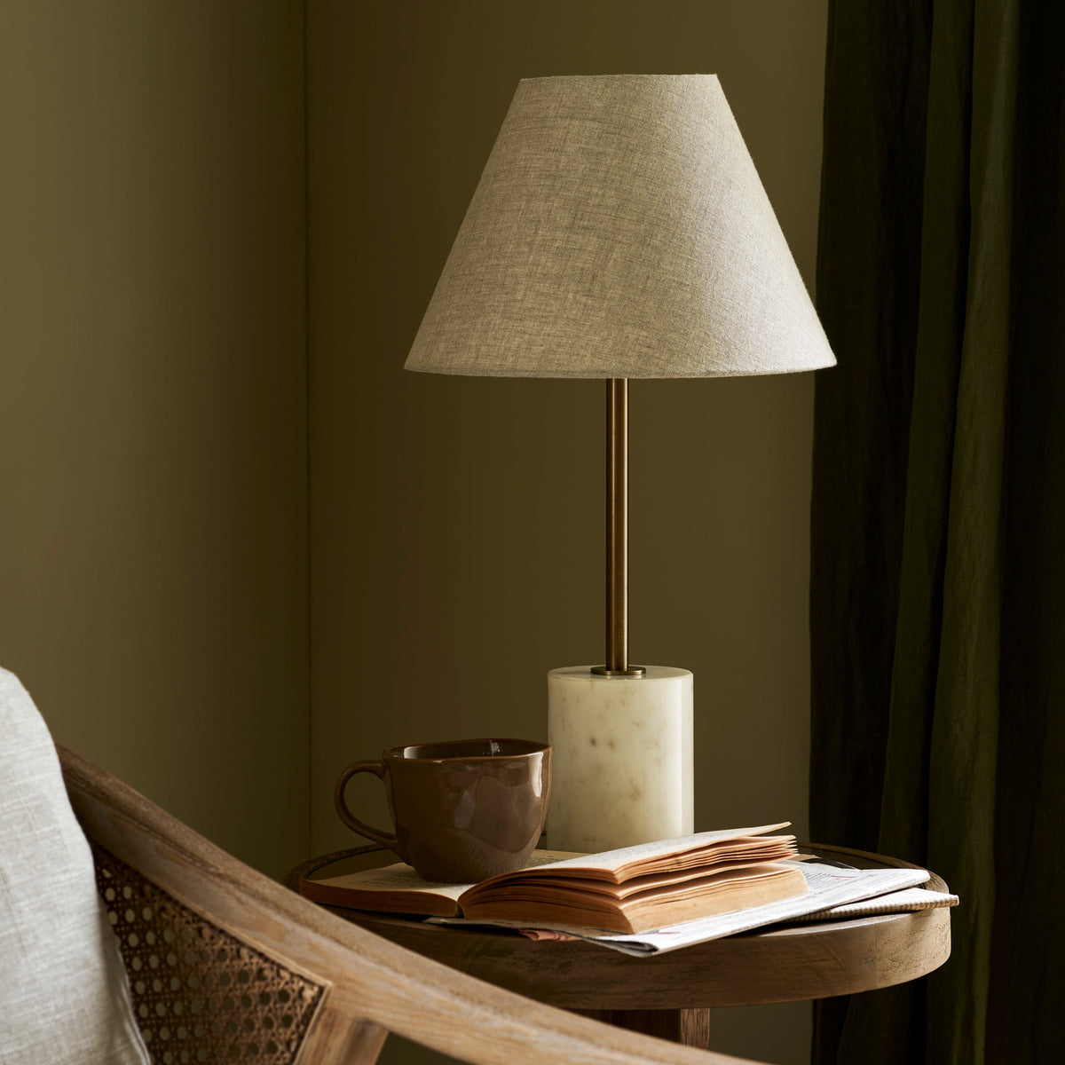 Winsley brass and marble table lamp with natural linen shade on a wooden table with a cup and books