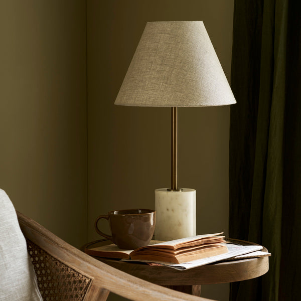 Winsley brass and marble table lamp with natural linen shade on a wooden table with a cup and books