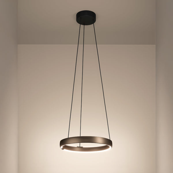 Modern circular pendant light with a bronze finish, emitting a warm glow, suspended from a black ceiling mount.