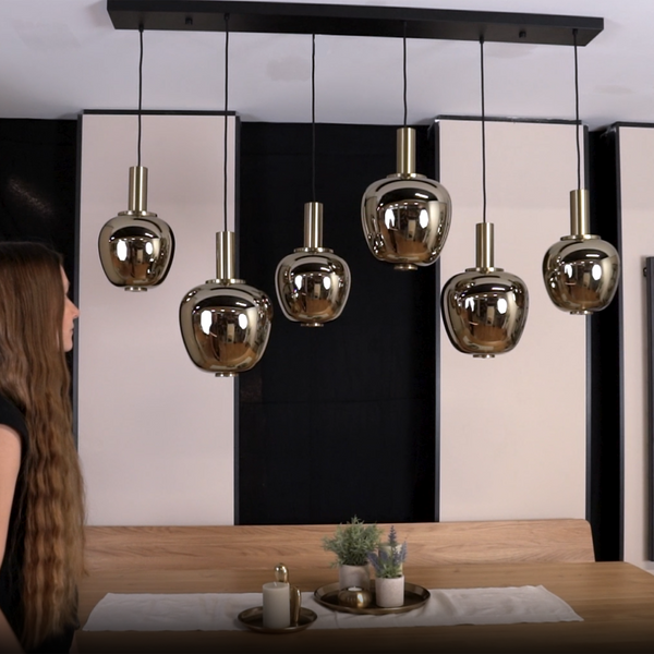 A woman stands next to a wooden dining table with six gold pendant lights hanging above it.