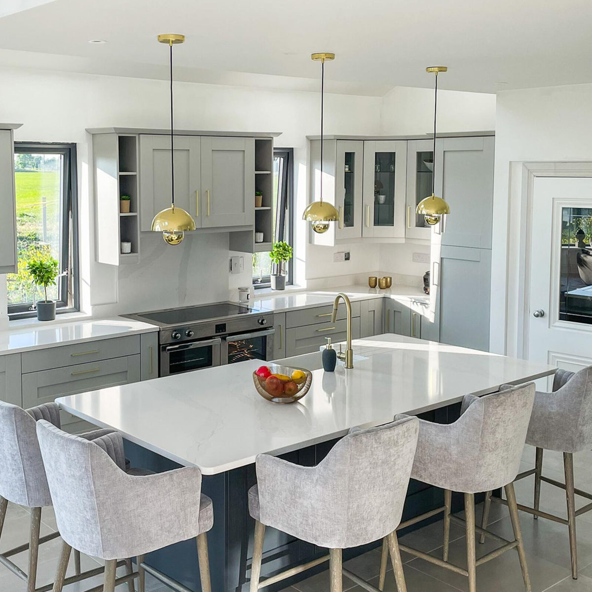 Modern kitchen with grey cabinets white countertops and gold pendant lights over a central island with grey bar stools
