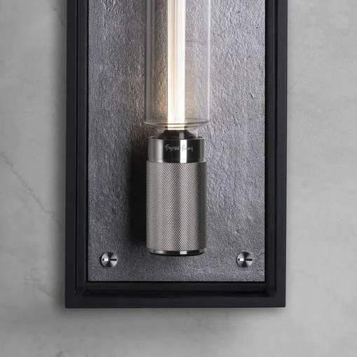 Close up of a black framed wall light with a textured grey backplate and a cylindrical glass shade