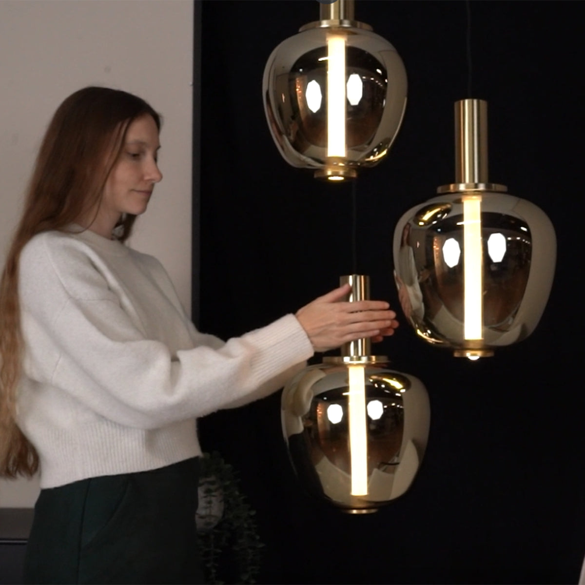 Woman adjusting a hanging gold pendant light with a glowing vertical bulb.