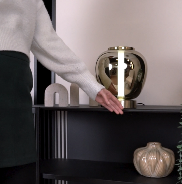 Woman in white sweater and dark skirt reaching for a gold lamp on a black shelf with decorative objects.