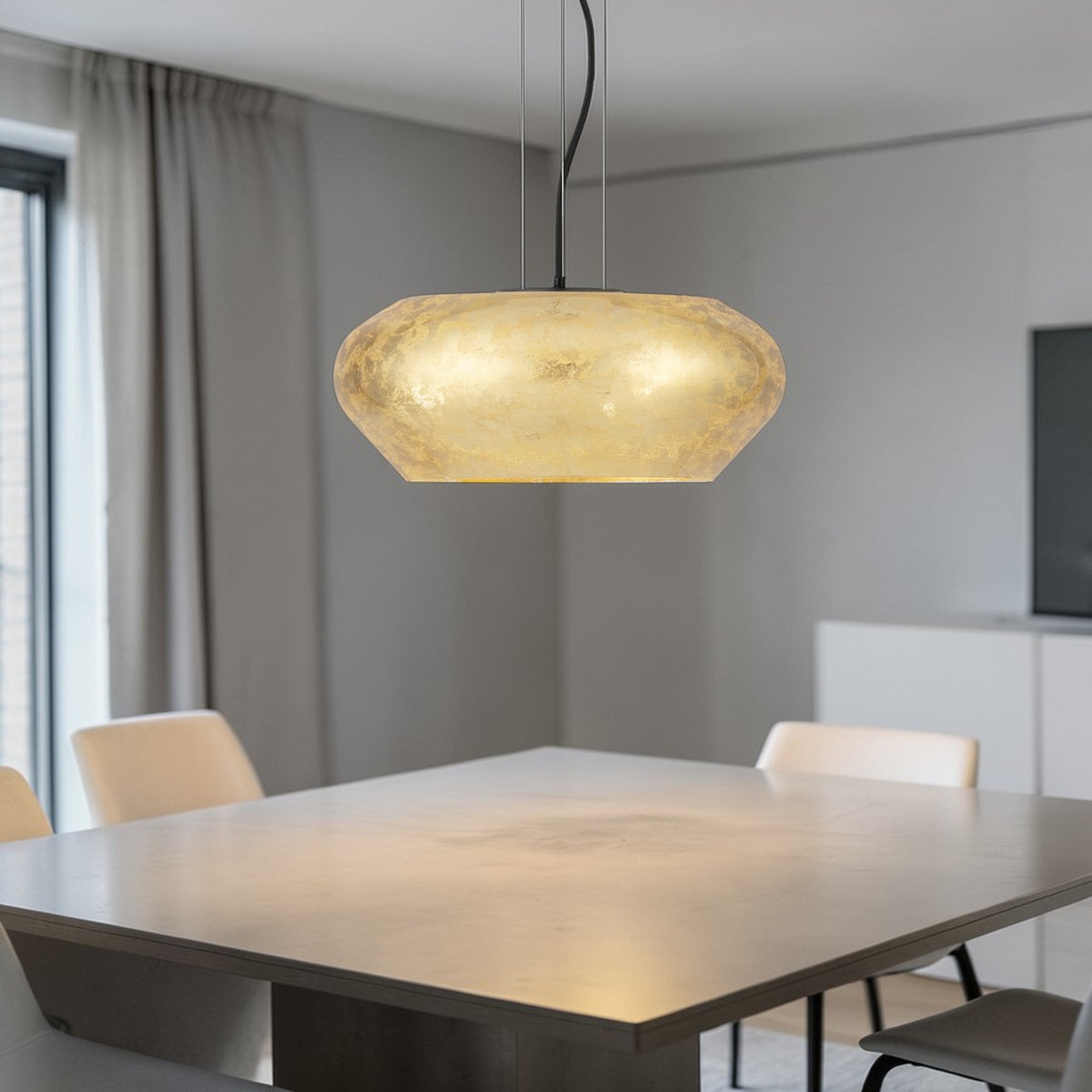 A modern dining room with a large, rectangular wooden table and light-colored chairs. A golden, textured pendant light hangs above the table, illuminating the space with a warm glow. The room features light grey walls and curtains, with a window visible on the left.
